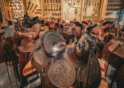 Interior of a western saddlery shop with elaborately tooled leather saddles on display and wall-mounted knives, rope, and gear in the background