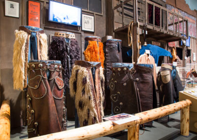 Row of leather chaps with fringed leather skirts and metal studs on display against a wooden wall in a western-themed exhibit.