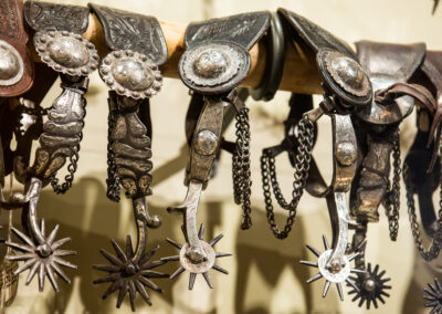 Decorative silver conchos and starburst spurs hanging on a row of leather western saddles.