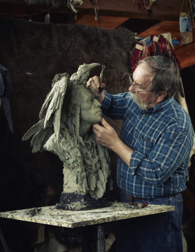 Adult man sculpting a large gray clay bust on a stand in a woodshop/studio.