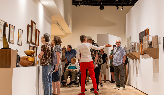 People tour a bright art gallery, viewing framed works on the walls and wooden sculptures on display pedestals.