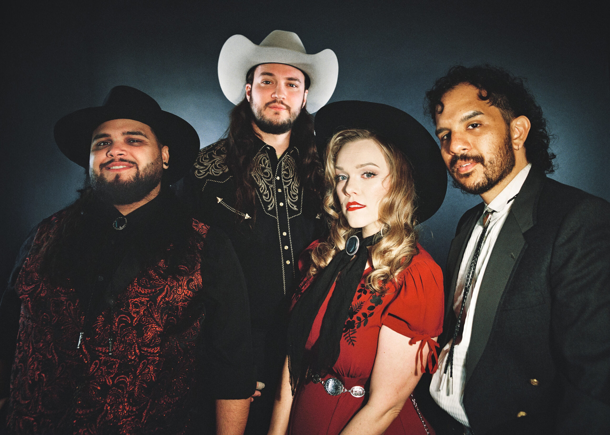 Group of four adults in western outfits posing together, two men wearing cowboy hats (one white, one black) and a woman in a red dress, against a dark background to the left.