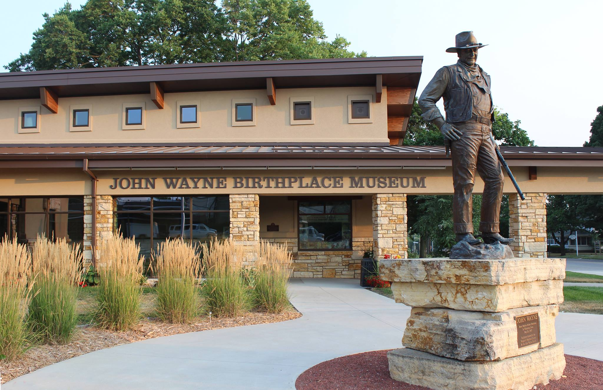 Bronze statue of a cowboy standing with a rifle beside the John Wayne Birthplace Museum building and sign in front of it